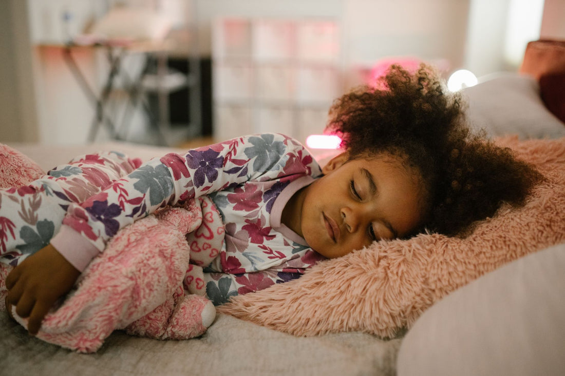 A young girl in floral pyjamas sleeping soundly on a soft pink blanket, with warm fairy lights glowing behind her